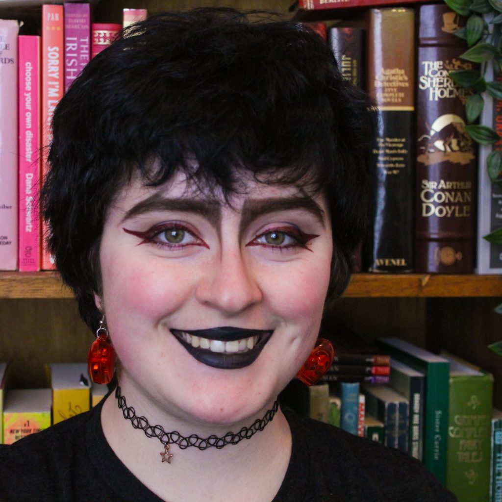 Photo of Anna Godfrey - person with short, wavy black hair, black lipstick and eye makeup, wearing a lace choker and a black shirt, standing in front of a bookshelf. 