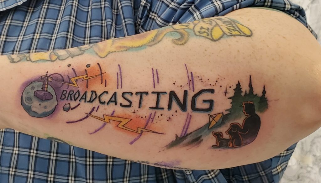 Photograph of the outer-side of somebody's forearm, showing a tattoo from their wrist to their elbow. The Moonbase Theta tower sits on the moon, sending out signal waves decorated with lightning bolts and stars to the silhouette of a person sitting with two dogs amongst the trees, next to a satellite dish. Text across the signal waves reads "BROADCASTING."